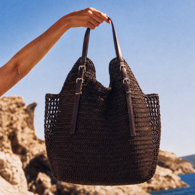 Brown woven handbag held by a person against a clear blue sky and rocky landscape.